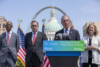Michael Bloomberg speaks at a podium with Andrew Martin and Lyda Krewson behind him